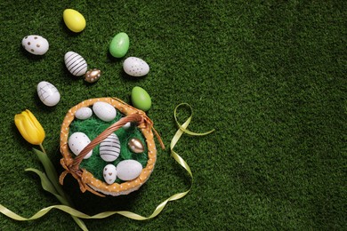 Photo of Easter basket with painted eggs, tulip and ribbon on green grass, flat lay. Space for text