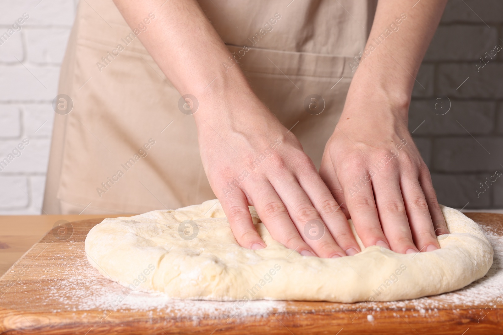 Photo of Woman kneading dough at wooden table near white brick wall, closeup