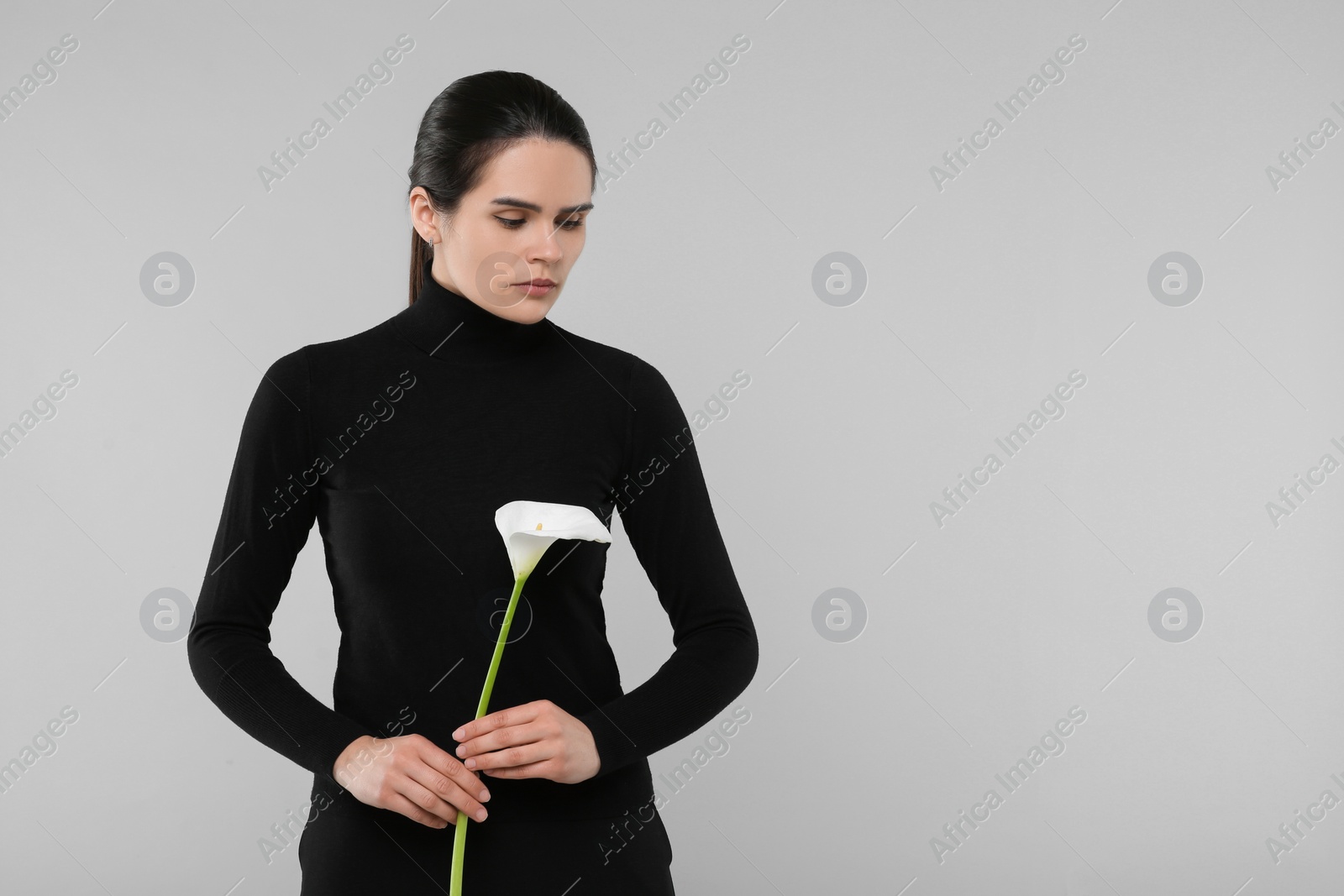 Photo of Sad woman with calla lily flower on light grey background, space for text. Funeral ceremony