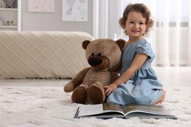 Photo of Cute little girl with teddy bear and book on floor at home