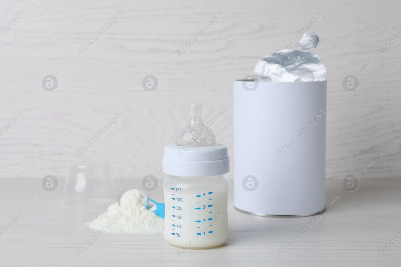 Photo of Blank can of powdered infant formula with scoop and feeding bottle on white wooden table. Baby milk