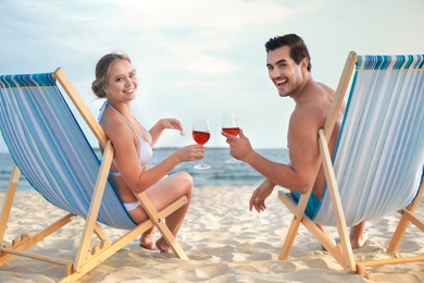 Happy young couple with glasses of wine sitting on deck chairs at sea beach