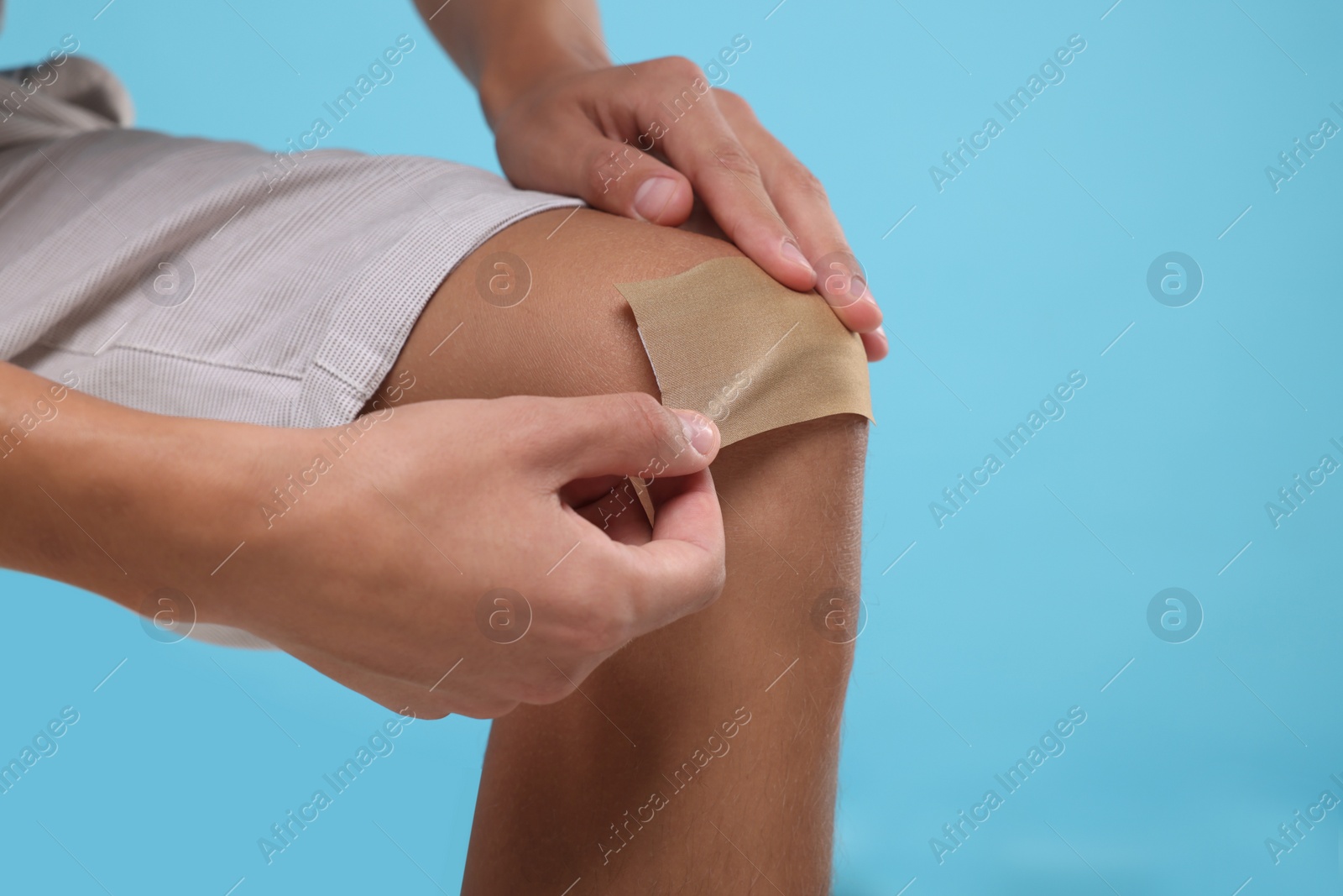 Photo of Man putting sticking plaster onto knee on light blue background, closeup. Space for text