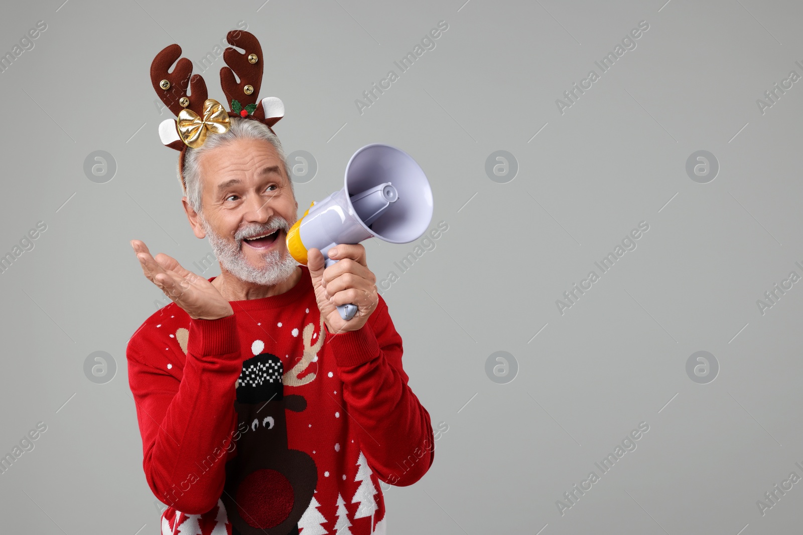 Photo of Senior man in Christmas sweater and reindeer headband shouting in megaphone on grey background. Space for text