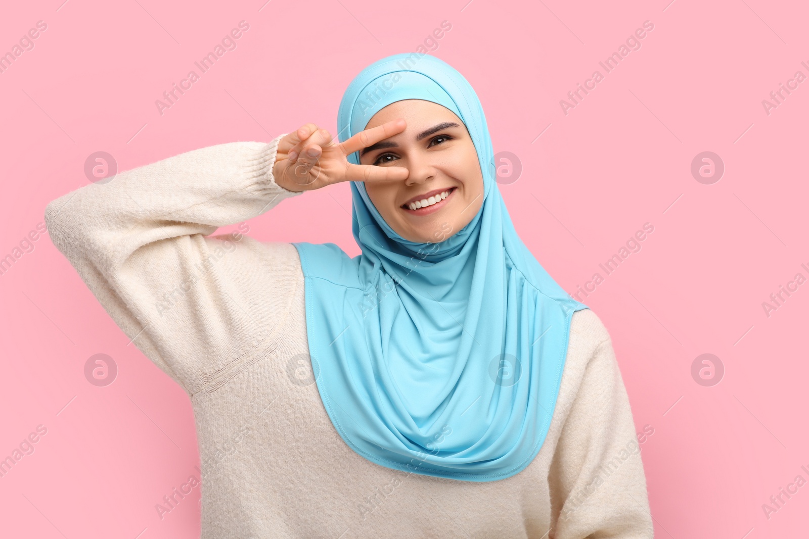 Photo of Muslim woman in hijab showing V-sign on pink background