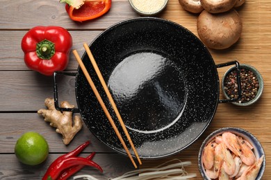 Black wok and chopsticks surrounded by products on color wooden table, flat lay