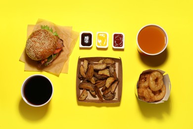 Photo of Tasty burger, potato wedges, fried onion rings, different sauces and refreshing drinks on yellow background, flat lay. Fast food