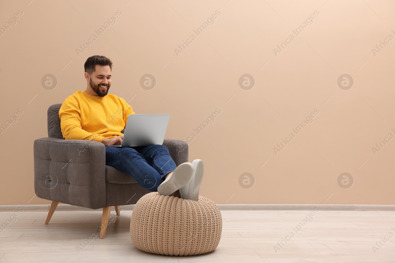 Photo of Handsome man with laptop sitting in armchair near beige wall indoors, space for text