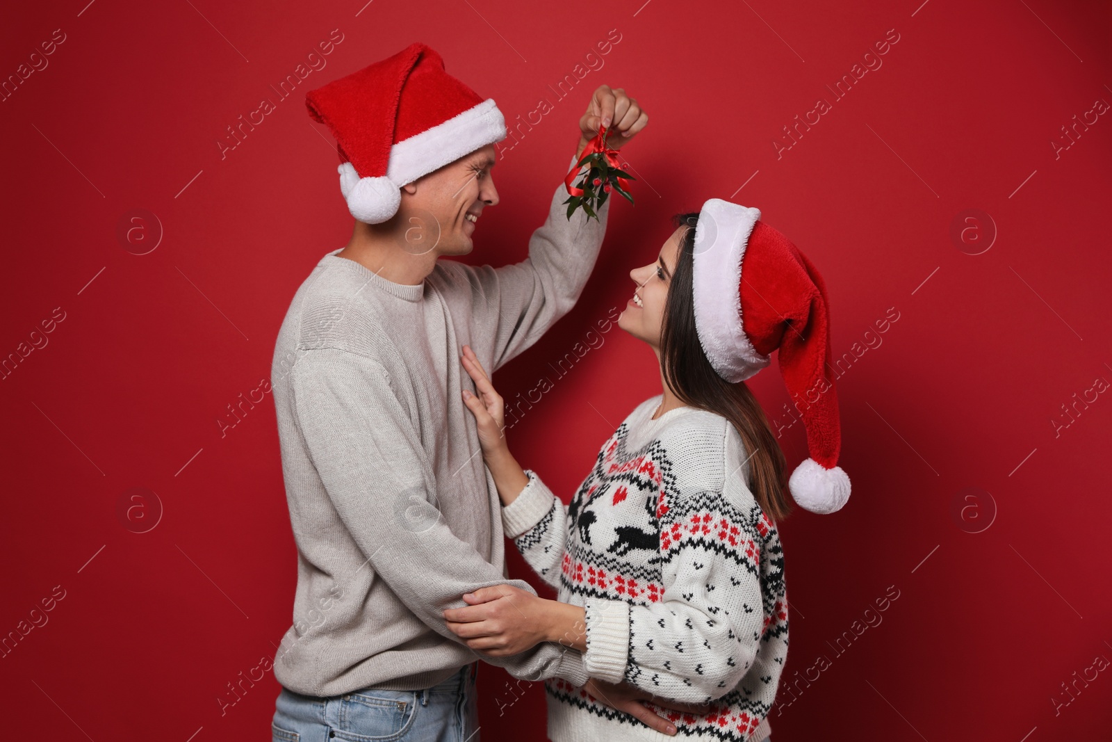 Photo of Happy couple in Santa hats standing under mistletoe bunch on red background