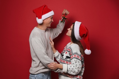 Happy couple in Santa hats standing under mistletoe bunch on red background