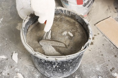 Worker with trowel mixing cement in bucket indoors, closeup