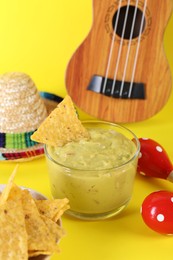 Delicious guacamole with nachos chips, Mexican sombrero hat, ukulele and maracas on yellow background