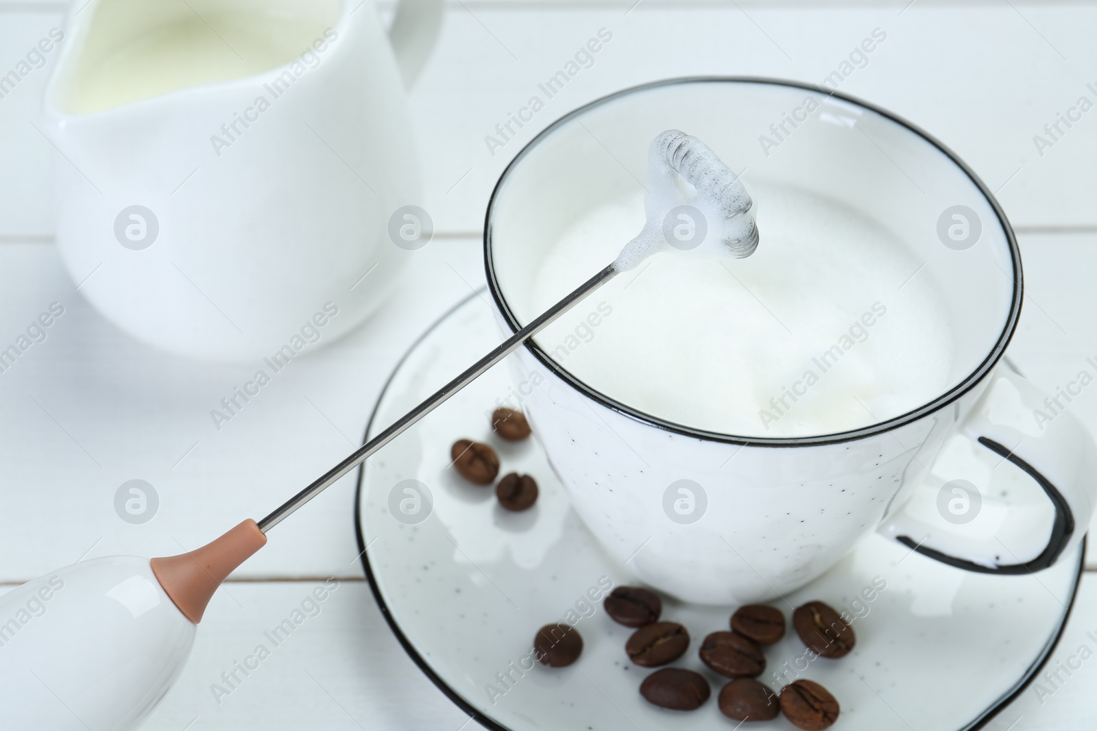 Photo of Mini mixer (milk frother), whipped milk and coffee beans on white wooden table, closeup