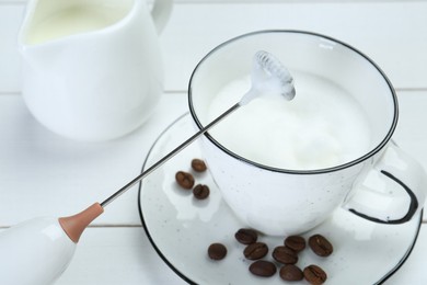 Mini mixer (milk frother), whipped milk and coffee beans on white wooden table, closeup