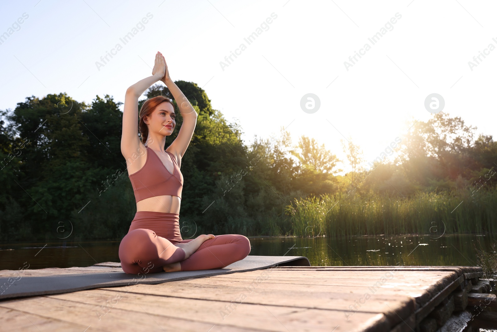 Photo of Beautiful woman practicing Padmasana on yoga mat on wooden pier near pond, space for text. Lotus pose