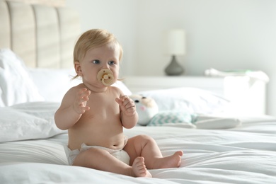 Cute little baby in diaper with pacifier sitting on bed at home. Space for text