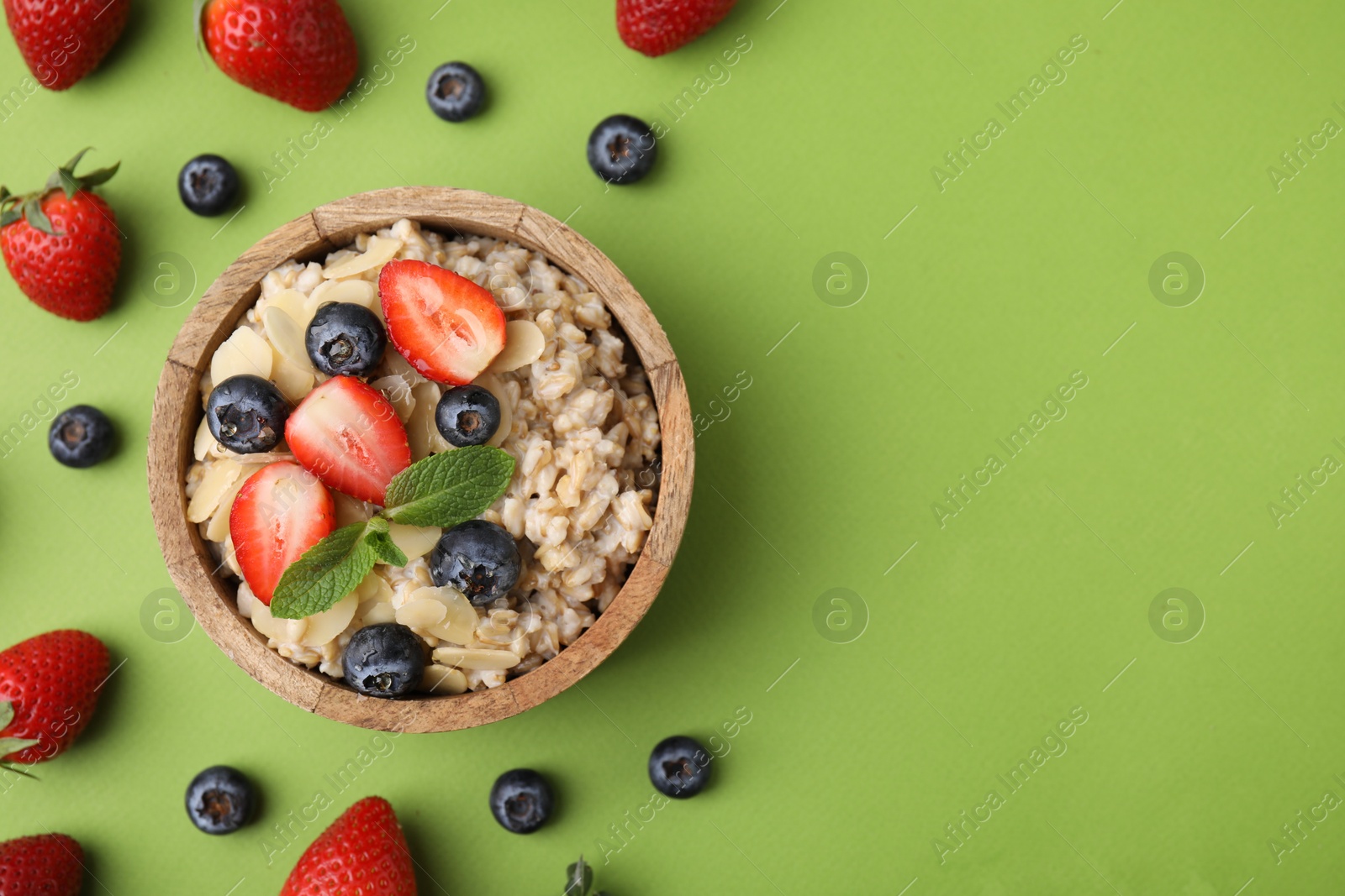 Photo of Tasty oatmeal with strawberries, blueberries and almond petals in bowl surrounded by fresh berries on green background, flat lay. Space for text