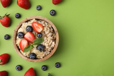 Photo of Tasty oatmeal with strawberries, blueberries and almond petals in bowl surrounded by fresh berries on green background, flat lay. Space for text