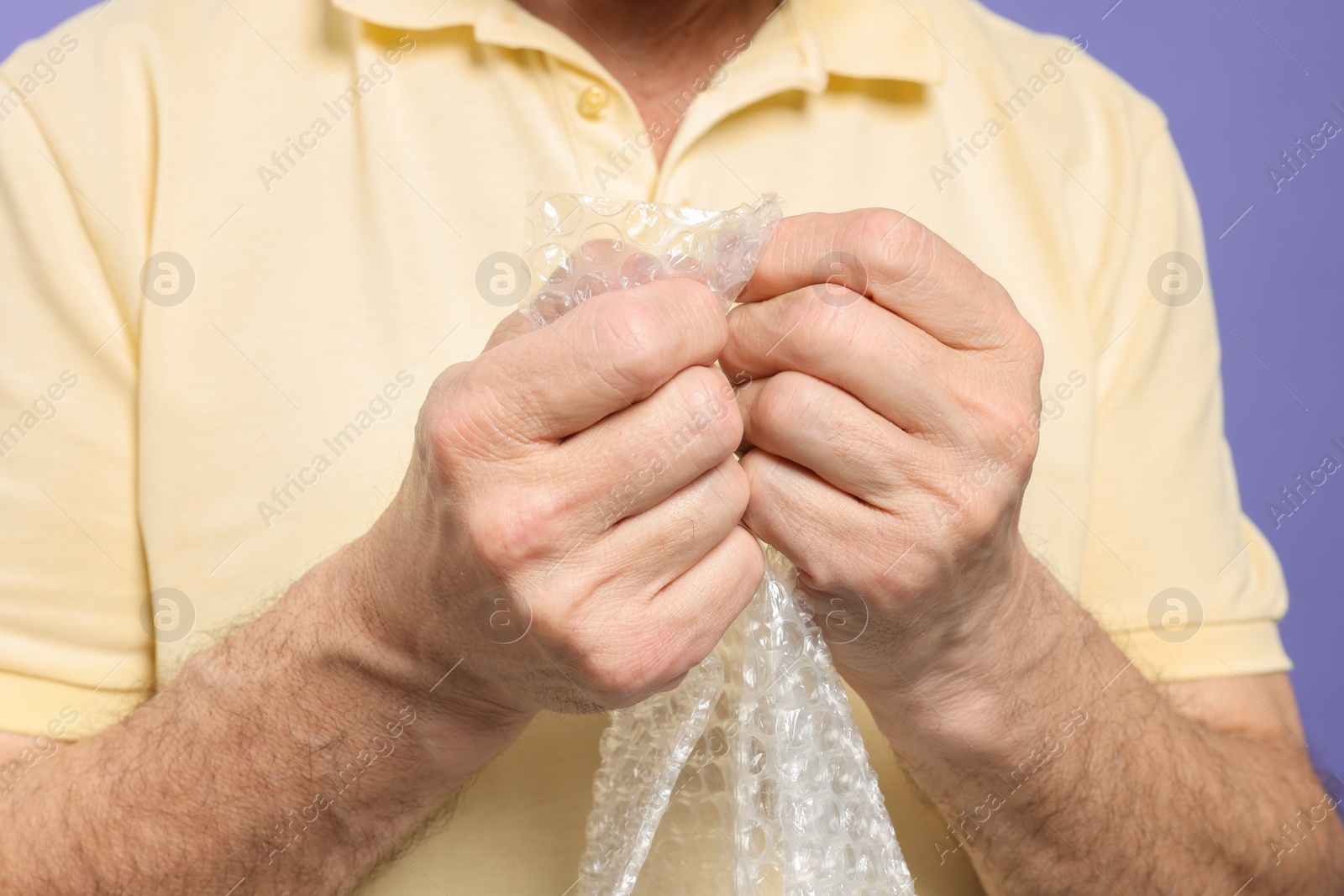 Photo of Senior man popping bubble wrap on light purple background, closeup. Stress relief