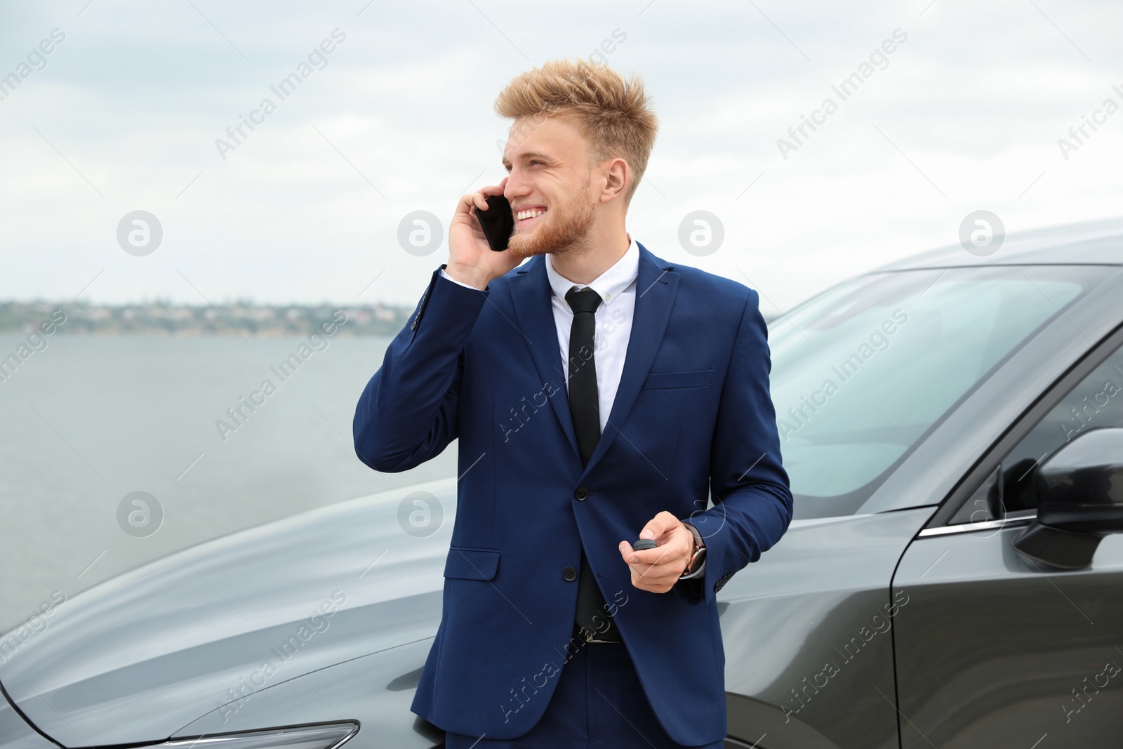 Photo of Young businessman with key talking on phone near car outdoors