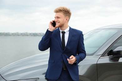 Young businessman with key talking on phone near car outdoors
