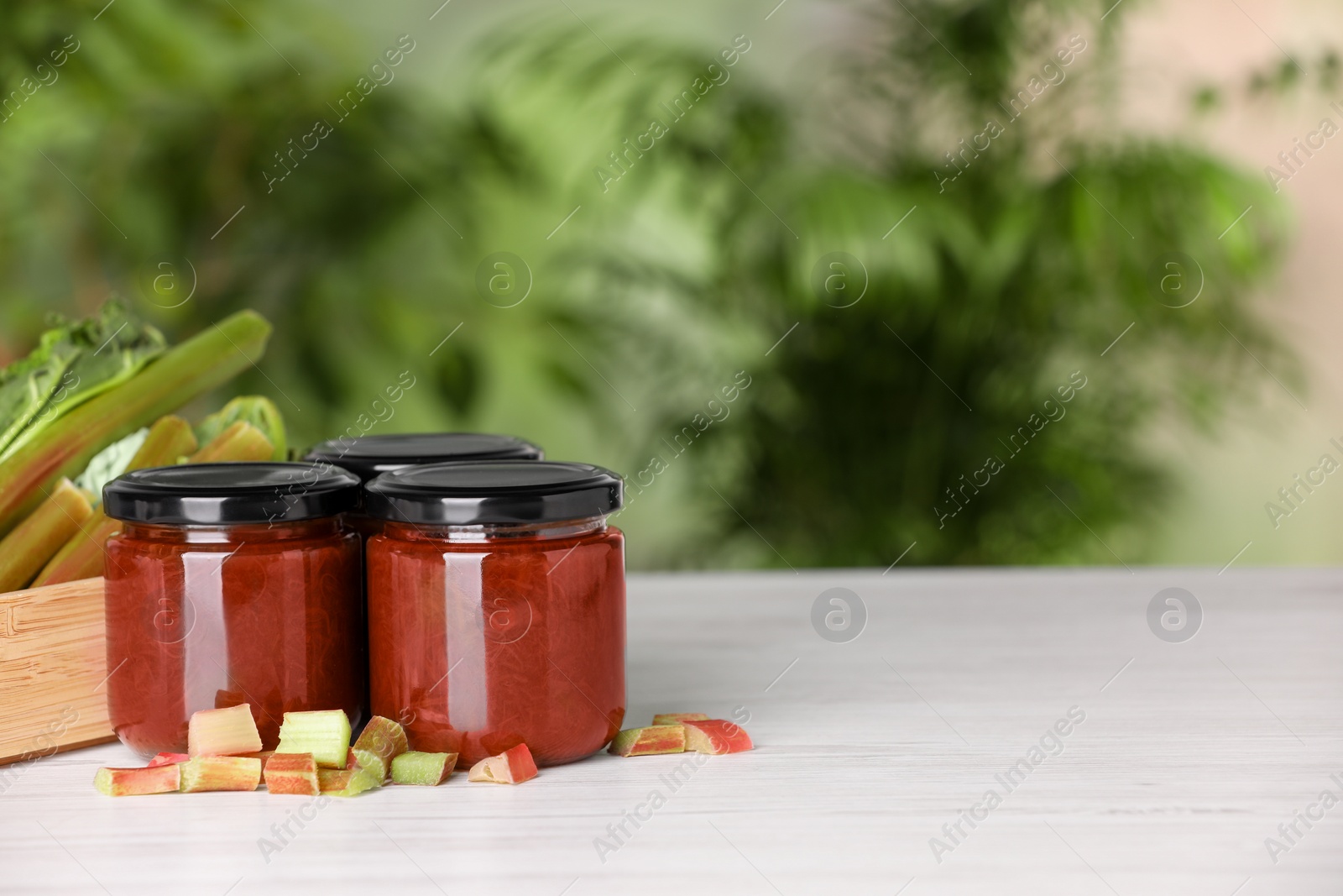 Photo of Jars of tasty rhubarb jam and stalks on white wooden table, space for text