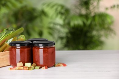 Photo of Jars of tasty rhubarb jam and stalks on white wooden table, space for text