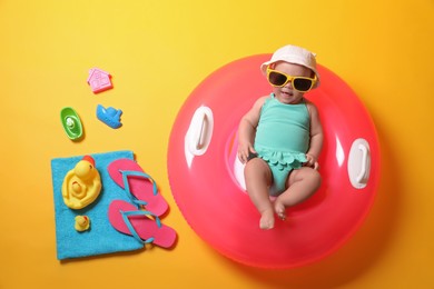 Photo of Cute little baby in sunglasses with inflatable ring and beach accessories on yellow background, top view