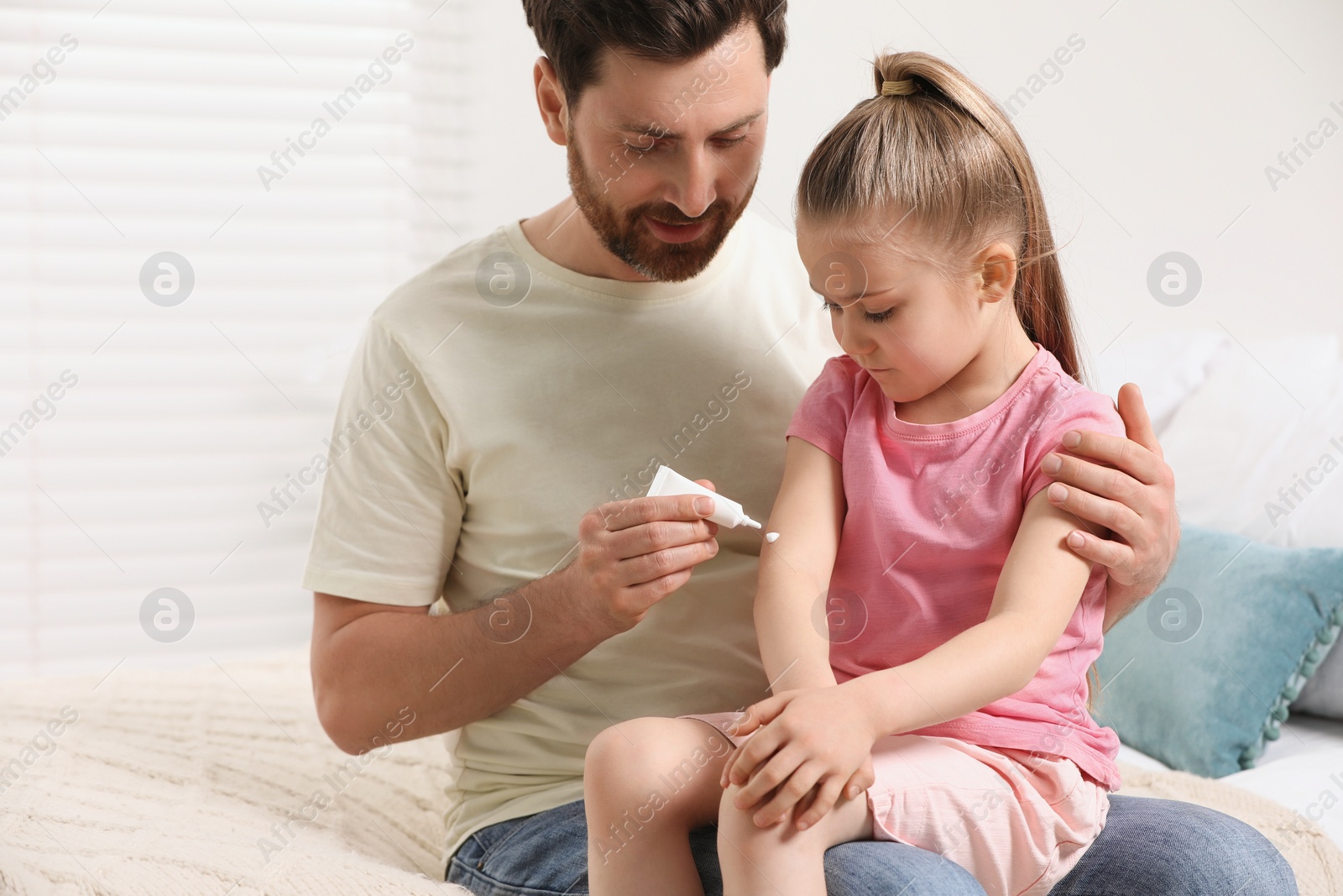 Photo of Father applying ointment onto his daughter's arm on bed indoors