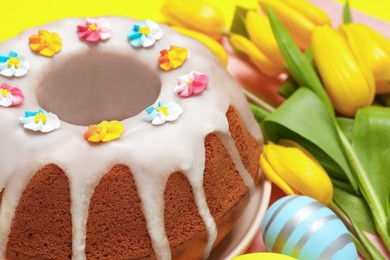 Photo of Delicious Easter cake decorated with sprinkles near beautiful tulips and painted egg on yellow background, closeup