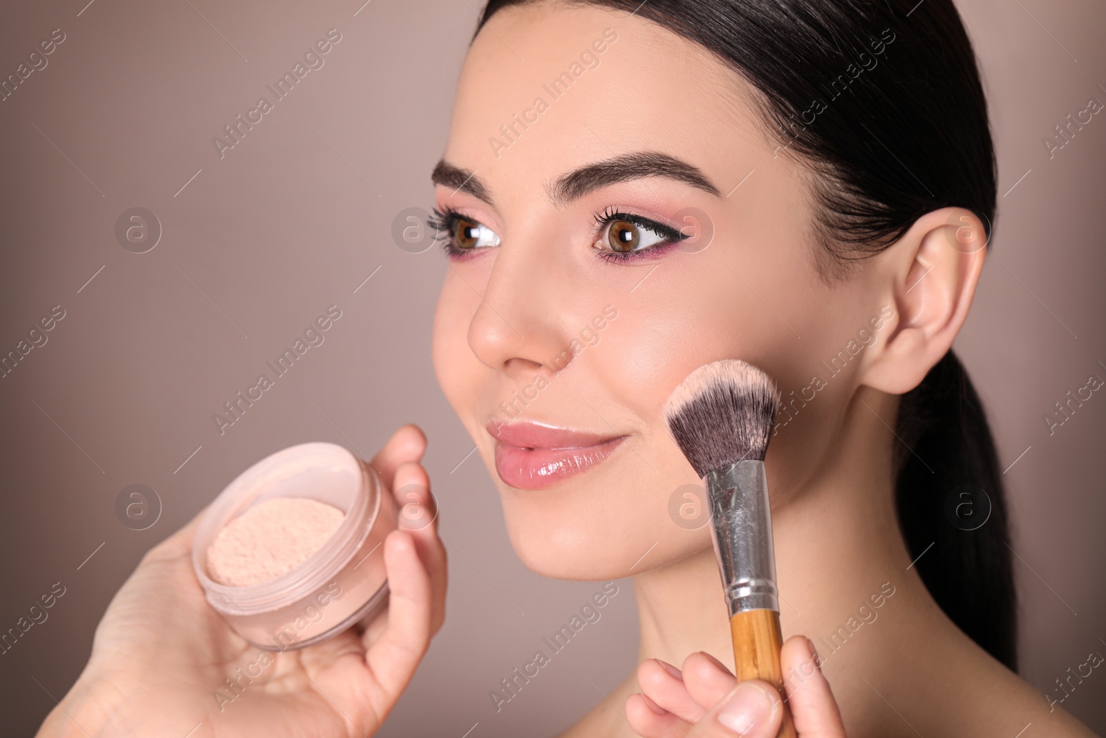 Photo of Professional makeup artist applying powder onto beautiful young woman's face with brush on dusty rose background, closeup