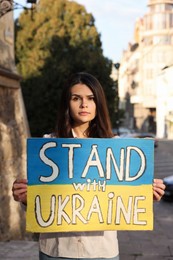 Photo of Sad woman holding poster in colors of national flag and words Stand with Ukraine on city street