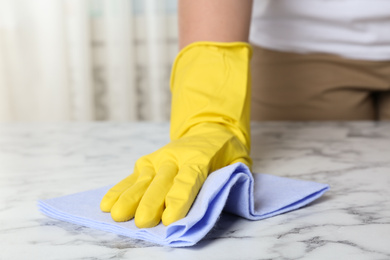 Photo of Woman in gloves wiping white marble table with rag indoors, closeup