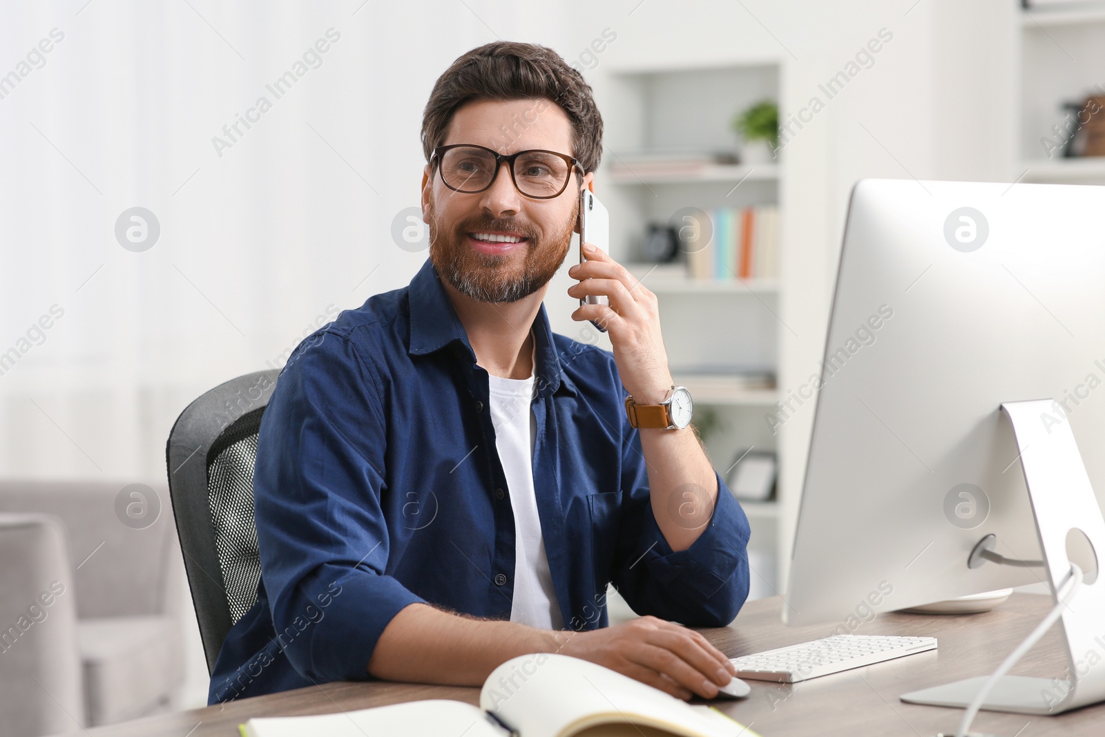 Photo of Home workplace. Happy man talking on smartphone while working with computer at wooden desk in room