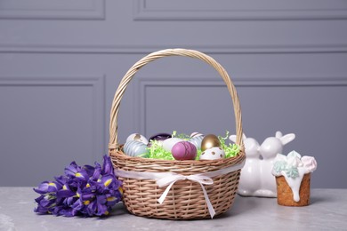 Photo of Easter basket with many painted eggs near tasty cake, flowers and figure of rabbits on grey textured table