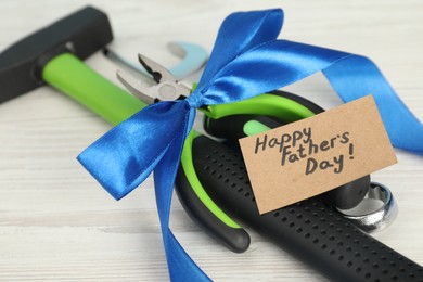 Photo of Happy Father's Day. Greeting card and set of tools tied with ribbon on light wooden table, closeup
