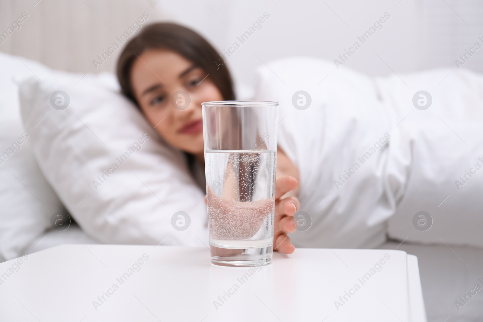 Photo of Young woman taking glass of water from nightstand at home, focus on hand