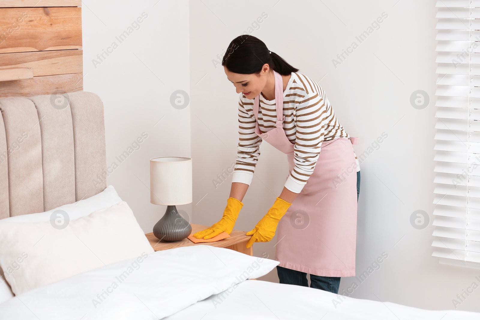 Photo of Young chambermaid wiping dust from nightstand in bedroom