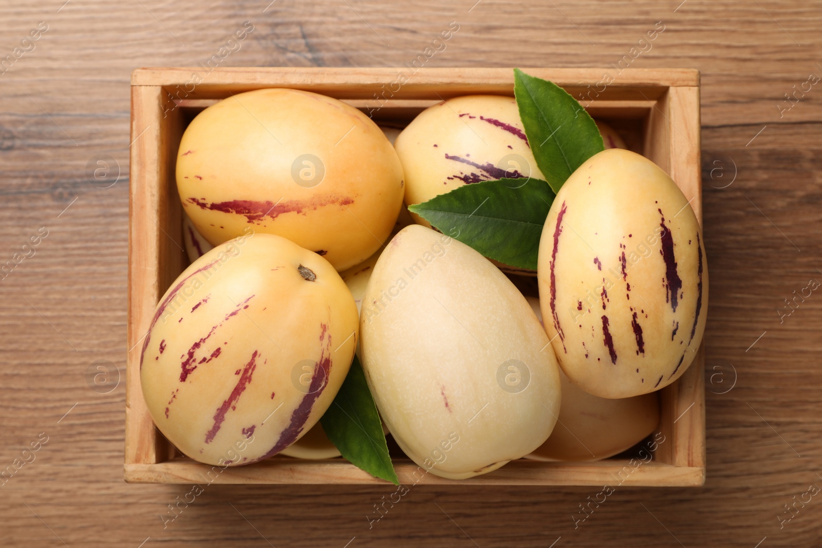 Photo of Fresh ripe pepino melons with green leaves in crate on wooden table, top view