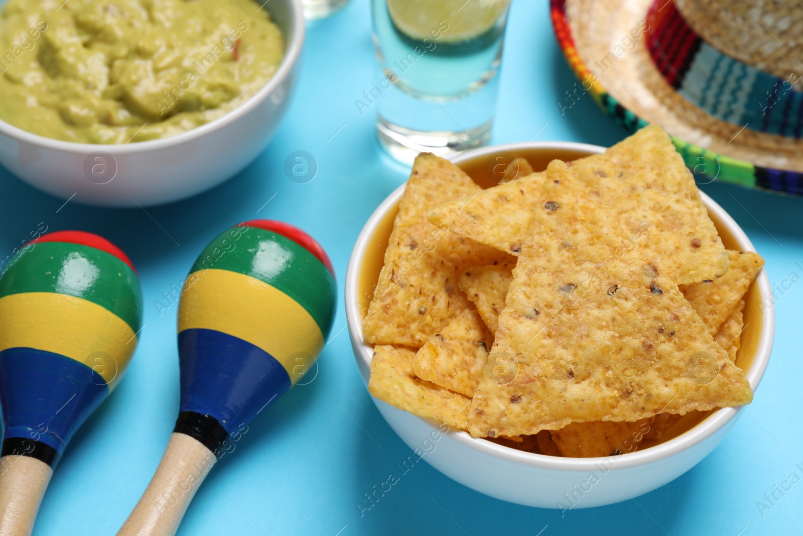 Photo of Nachos chips, Mexican sombrero hat, maracas, guacamole and tequila on light blue background, closeup