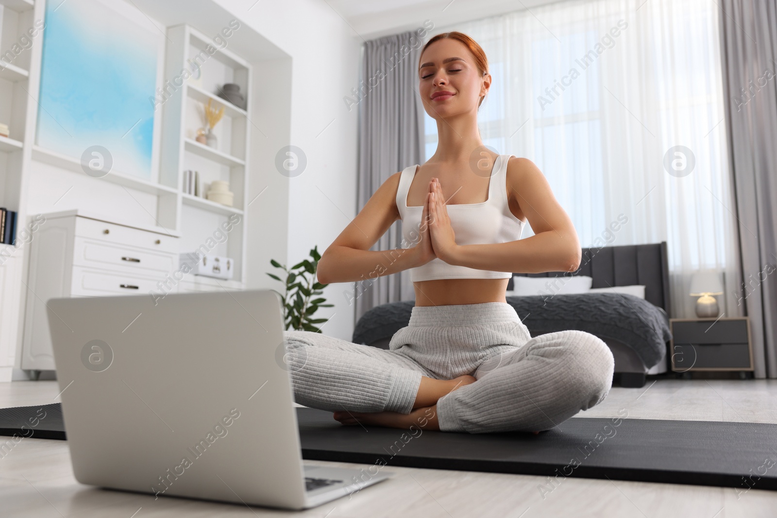 Photo of Beautiful young woman practicing Padmasana with laptop on yoga mat at home, low angle view. Lotus pose