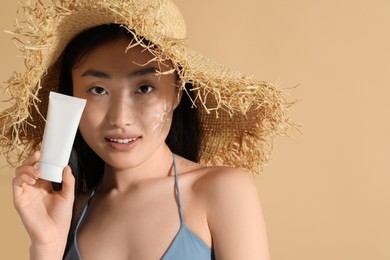 Photo of Beautiful young woman in straw hat with sunscreen on her face holding sun protection cream against beige background, space for text