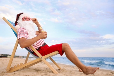 Photo of Santa Claus with cocktail relaxing in chair on beach, space for text. Christmas vacation