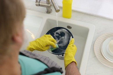 Man in protective gloves washing plate above sink indoors, closeup