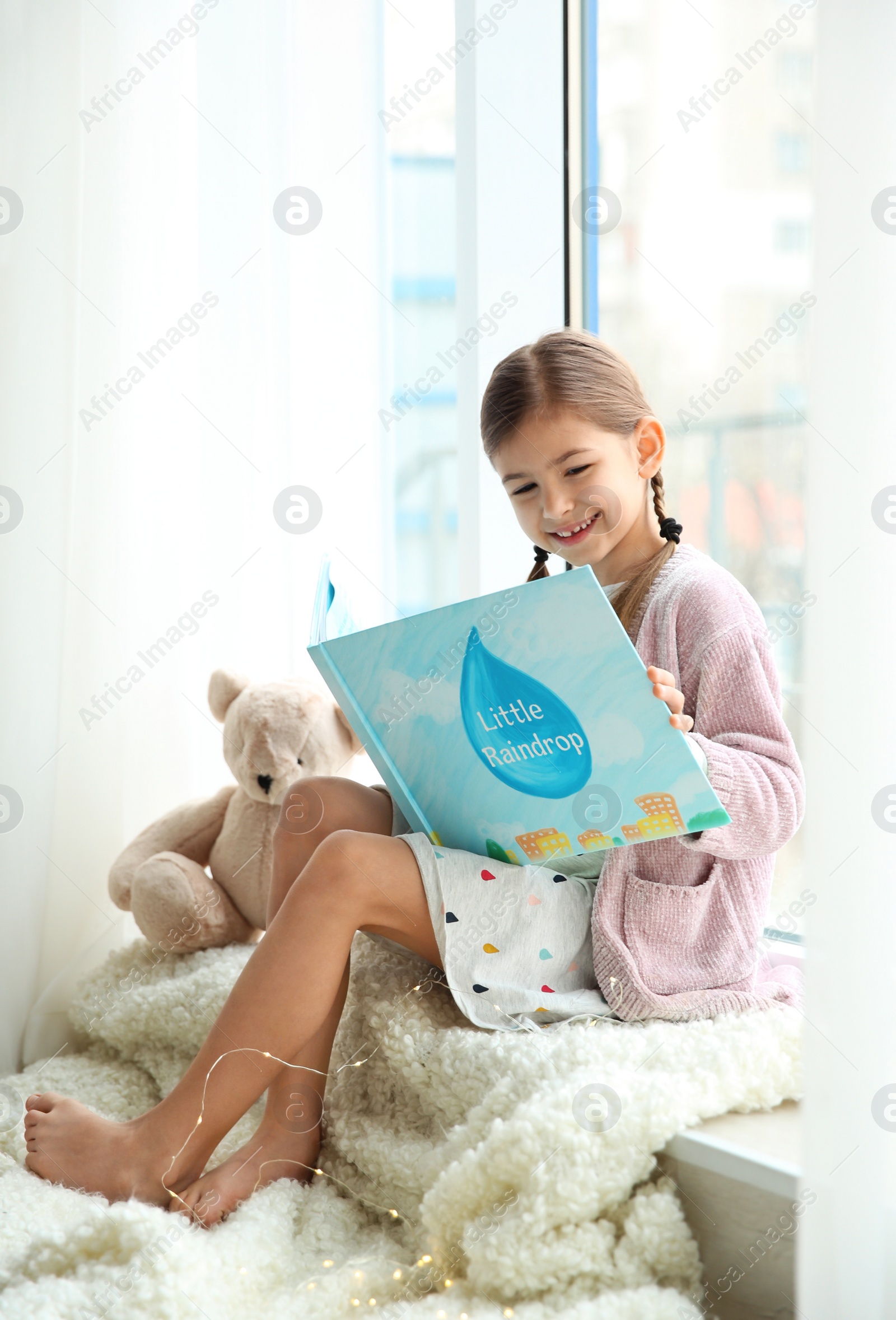 Photo of Cute little girl reading book near window at home