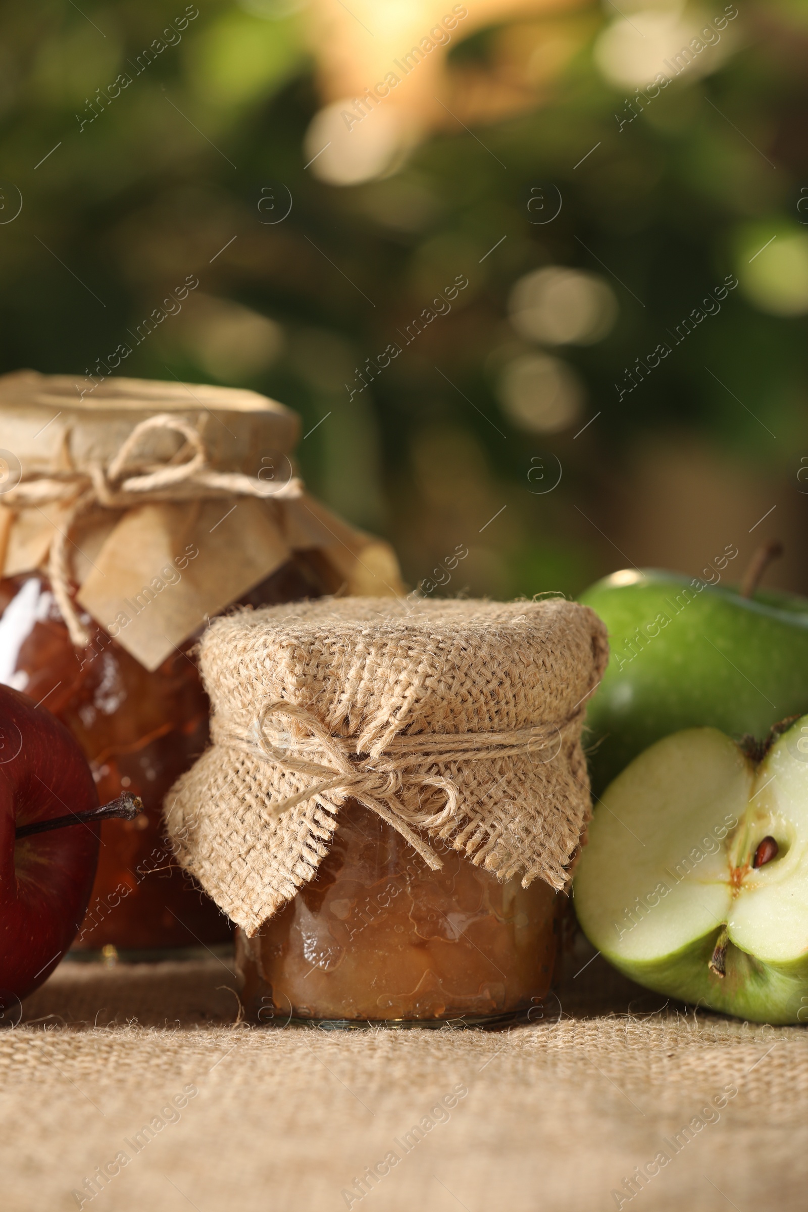Photo of Delicious apple jam and fresh fruits on burlap against blurred background, closeup. Space for text