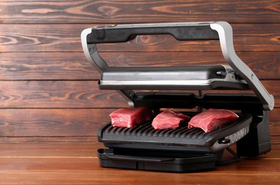 Photo of Electric grill with raw meat on wooden table. Space for text