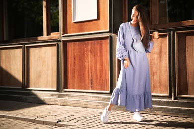 Photo of Beautiful young woman in stylish blue dress with handbag and cup of coffee near wooden building outdoors