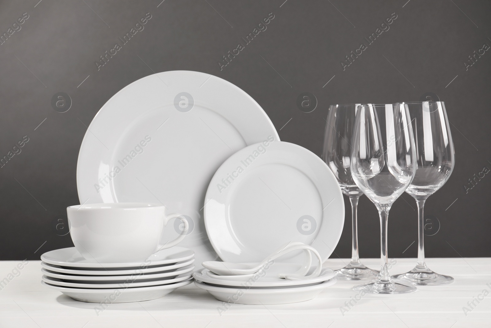 Photo of Set of clean dishware and glasses on white wooden table against grey background