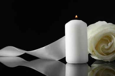 Burning candle, white rose and ribbon on black mirror surface in darkness, closeup with space for text. Funeral symbols